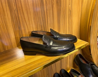 Op de afbeelding: Een paar zwarte leren penny loafers op een houten plank. De schoenen hebben een klassiek ontwerp met een ronde neus, een band over de wreef en een lage hak. De plank is gemaakt van lichtgekleurd hout.