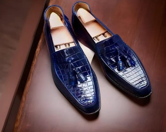 May include: A pair of blue leather loafers with a crocodile skin texture. Each shoe has a tassel and a wooden shoe tree inside. The shoes are displayed on a dark brown surface.