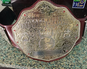Puede incluir: Un cinturón de campeonato de lucha libre dorado con las palabras "WORLD HEAVY WEIGHT WRESTLING CHAMPION" grabadas en la parte delantera. El cinturón tiene una correa de cuero burdeos y está decorado con joyas rojas.