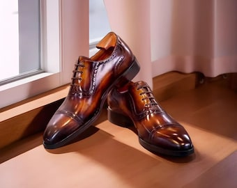 May include: A pair of brown leather dress shoes with a glossy finish. The shoes have a classic Oxford design with intricate detailing and a lace-up closure. The shoes are displayed near a window.
