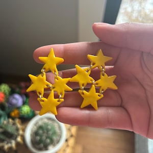 May include: A pair of bright yellow star-shaped earrings. Each earring features three connected stars, suspended from a gold-colored chain. The earrings are held up by a hand, with a blurred background of plants and a window.