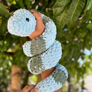 May include: A crocheted snake toy in light blue and green. The snake is curled up and being held by a person's hands.