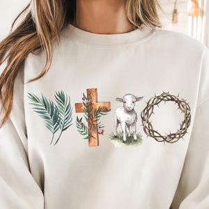 May include: Cream-colored sweatshirt featuring watercolor illustrations of a palm branch, a wooden cross adorned with greenery, a lamb, and a crown of thorns. The design is centered on the front of the long-sleeved top.