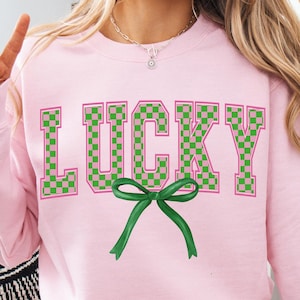 May include: A pink sweatshirt with the word "LUCKY" printed in green and pink checkered letters. A green bow is centered below the word.
