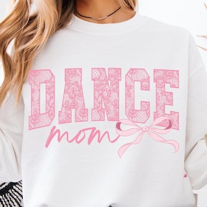 Op de afbeelding: Een wit sweatshirt met de tekst "DANCE mom" in roze met een roze strik. De tekst is versierd met een roze bloemenpatroon.