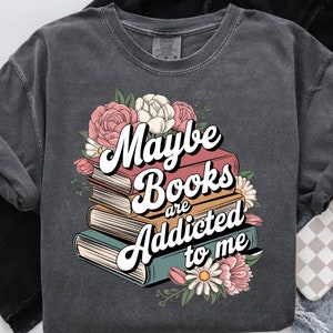May include: A dark gray t-shirt with a stack of books and flowers. The text on the shirt reads "Maybe books are addicted to me".