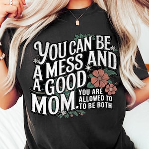 Może przedstawiać: Czarny T-shirt z białym nadrukiem, który głosi "You can be a mess and a good mom. You are allowed to be both." Nadruk jest otoczony małymi różowymi kwiatami.