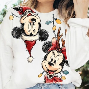 May include: White sweatshirt featuring a festive design with Mickey and Minnie Mouse. Mickey is wearing a Santa hat and Minnie has reindeer antlers. Both characters are adorned with string lights, creating a holiday theme.
