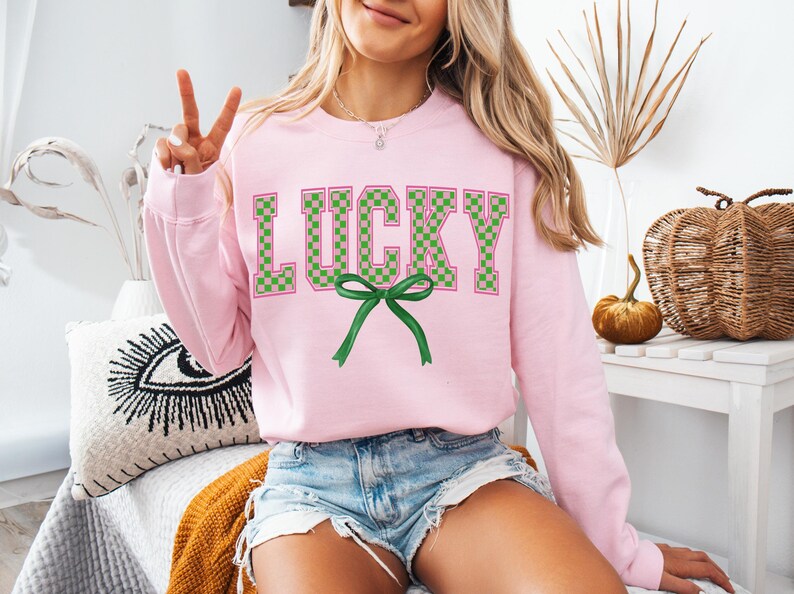 May include: A pink sweatshirt with the word "LUCKY" printed in green and pink checkered letters. A green bow is centered below the word.