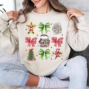 May include: Beige sweatshirt with a Christmas-themed design. The design includes images of a Santa Claus, bows, a Christmas tree, and a camper with a lightning bolt. The text on the sweatshirt reads "You Serious Clark?"
