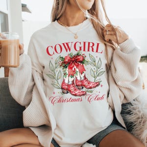 Op de afbeelding: Cr&egrave;mekleurige sweatshirt met de rode tekst "COWGIRL". Daaronder een afbeelding van rode cowboylaarzen met een rode strik en hulst. De woorden "Christmas Club" staan eronder. De sweatshirt heeft lange mouwen.