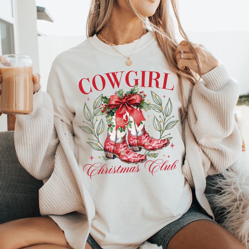 Op de afbeelding: Cr&egrave;mekleurige sweatshirt met de rode tekst "COWGIRL". Daaronder een afbeelding van rode cowboylaarzen met een rode strik en hulst. De woorden "Christmas Club" staan eronder. De sweatshirt heeft lange mouwen.