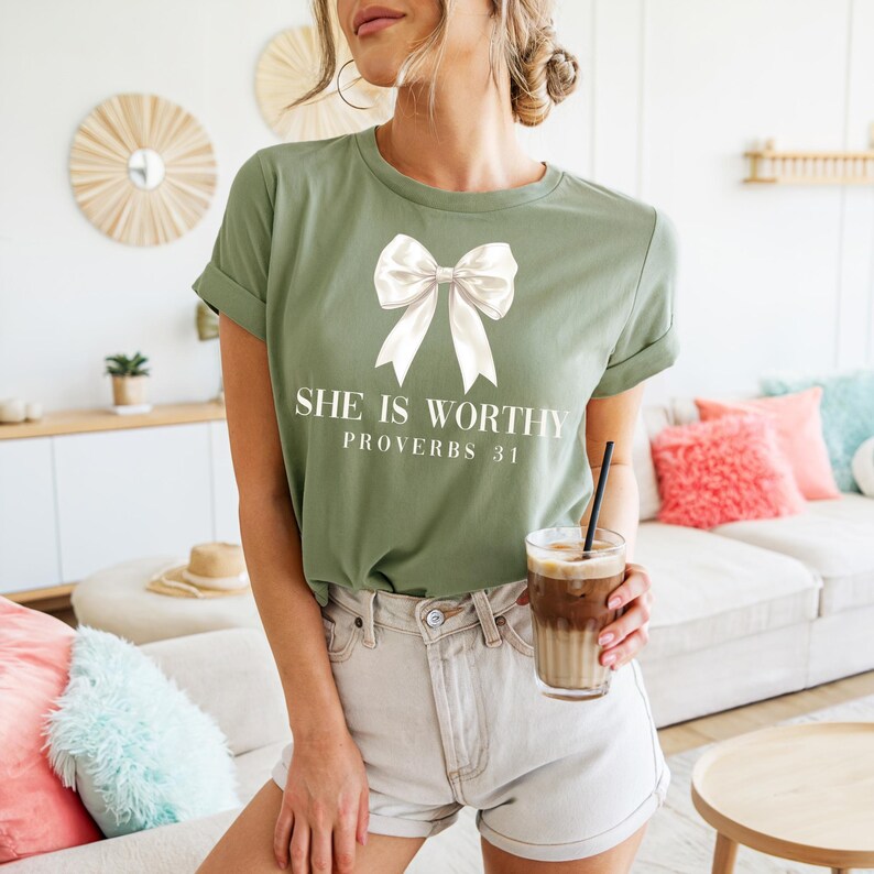May include: Olive green t-shirt with a white bow graphic and the text "SHE IS WORTHY PROVERBS 31". The shirt has rolled-up sleeves and is paired with light-colored shorts. A glass of iced coffee is held in the hand.
