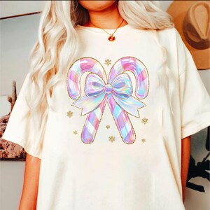 May include: Cream-colored t-shirt featuring a whimsical design of two candy canes and a bow in iridescent pastel colors. The candy canes have a plaid pattern and are outlined in gold glitter. Small gold snowflakes are scattered around the design.