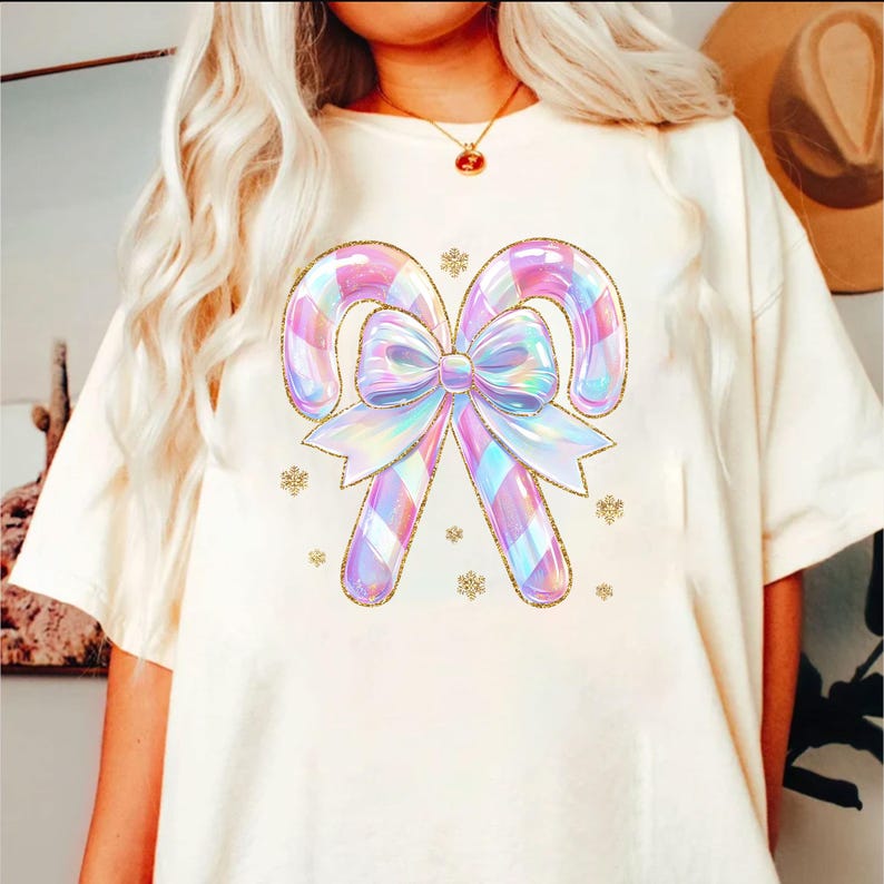 May include: Cream-colored t-shirt featuring a whimsical design of two candy canes and a bow in iridescent pastel colors. The candy canes have a plaid pattern and are outlined in gold glitter. Small gold snowflakes are scattered around the design.