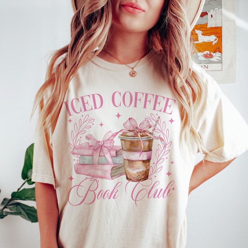 K&ouml;nnte beinhalten: Ein wei&szlig;es T-Shirt mit einem rosafarbenen Grafikdesign, auf dem "Iced Coffee Book Club" steht. Die Grafik zeigt einen Stapel B&uuml;cher mit einer rosafarbenen Schleife, eine Tasse Eiskaffee mit einer rosafarbenen Schleife und einem Strohhalm sowie rosafarbene Bl&auml;tter.