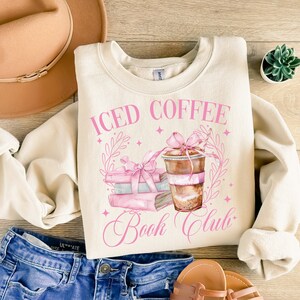 K&ouml;nnte beinhalten: Ein cremefarbener Pullover mit dem Text "ICED COFFEE BOOK CLUB" in rosa Schrift. Das Design zeigt einen Stapel B&uuml;cher mit einer rosa Schleife und eine Tasse Eiskaffee mit einer rosa Schleife.