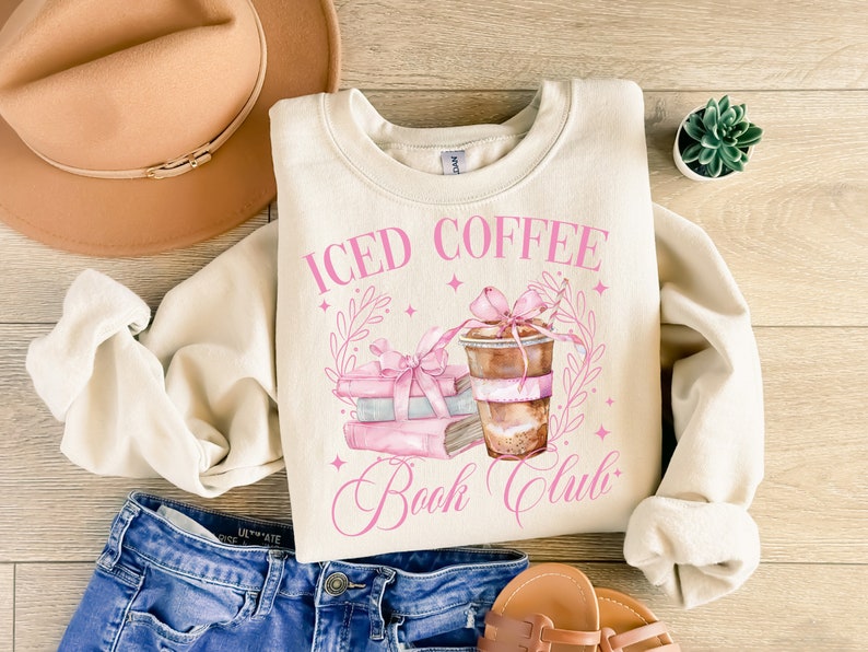 K&ouml;nnte beinhalten: Ein cremefarbener Pullover mit dem Text "ICED COFFEE BOOK CLUB" in rosa Schrift. Das Design zeigt einen Stapel B&uuml;cher mit einer rosa Schleife und eine Tasse Eiskaffee mit einer rosa Schleife.