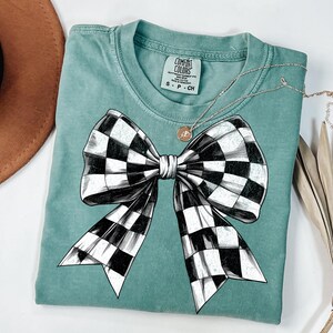 May include: A teal blue t-shirt featuring a black and white checked bow design. The shirt has a label that reads "COMFORT COLORS".