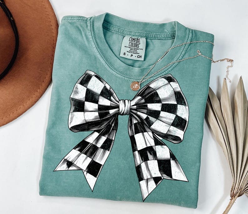 May include: A teal blue t-shirt featuring a black and white checked bow design. The shirt has a label that reads "COMFORT COLORS".