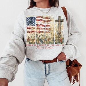 May include: A gray sweatshirt with a watercolor design of an American flag and a cross. The design is surrounded by flowers and the text "Where the Spirit of the Lord is There is Freedom".