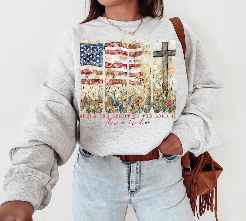 May include: A gray sweatshirt with a watercolor design of an American flag and a cross. The design is surrounded by flowers and the text "Where the Spirit of the Lord is There is Freedom".