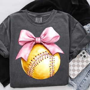 May include: A gray t-shirt with a pink bow on top of a yellow softball. The softball has white stitching.