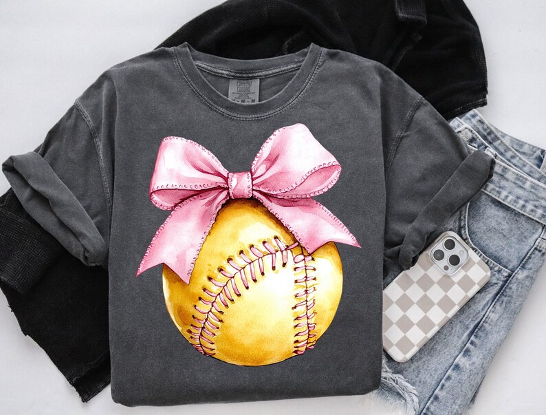 May include: A gray t-shirt with a pink bow on top of a yellow softball. The softball has white stitching.
