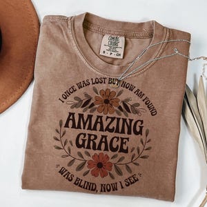 May include: A brown t-shirt with a floral design and the text "I once was lost but now am found Amazing Grace Was blind, now I see".