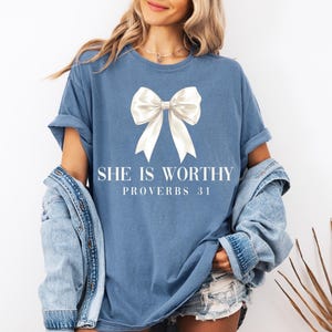 May include: A blue t-shirt with a large white bow graphic and the text "SHE IS WORTHY PROVERBS 31". The shirt has rolled-up sleeves and is paired with a denim jacket and shorts.
