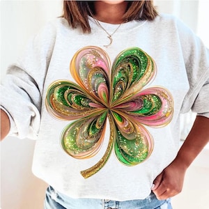 May include: A white sweatshirt featuring a green, gold and pink four-leaf clover design. The clover is made of swirling lines and glitter.