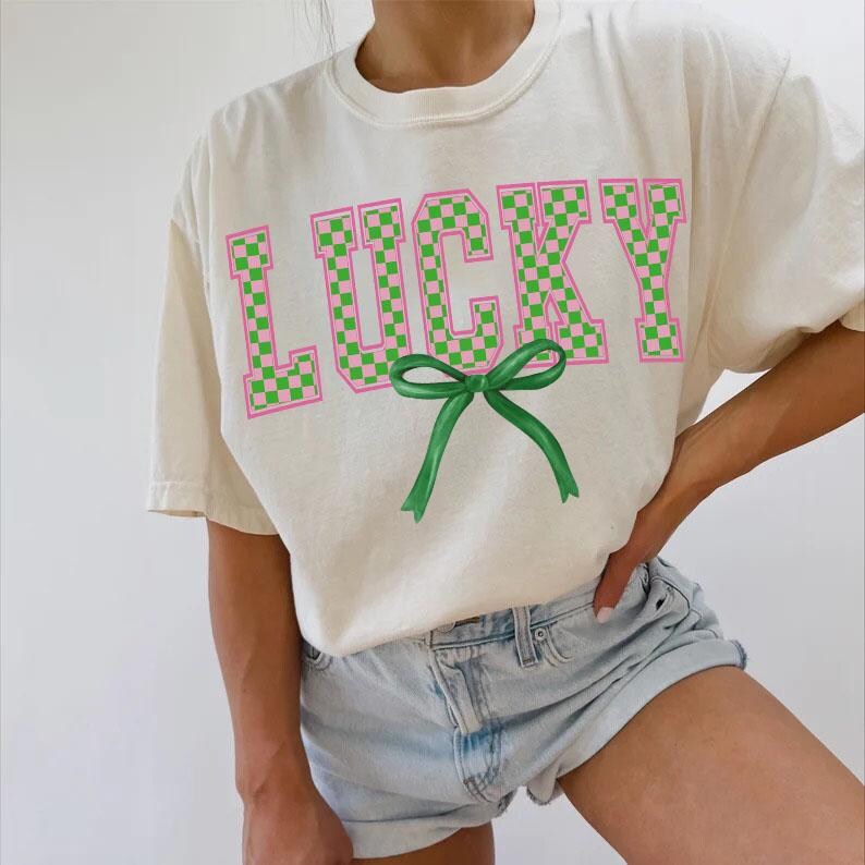 May include: A white t-shirt with the word "LUCKY" printed in green and pink checkered letters. A green bow is below the word.