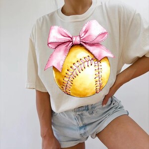 May include: A white t-shirt with a yellow softball and a pink bow on it. The softball has white stitching.