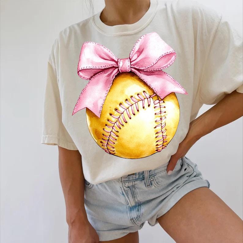 May include: A white t-shirt with a yellow softball and a pink bow on it. The softball has white stitching.