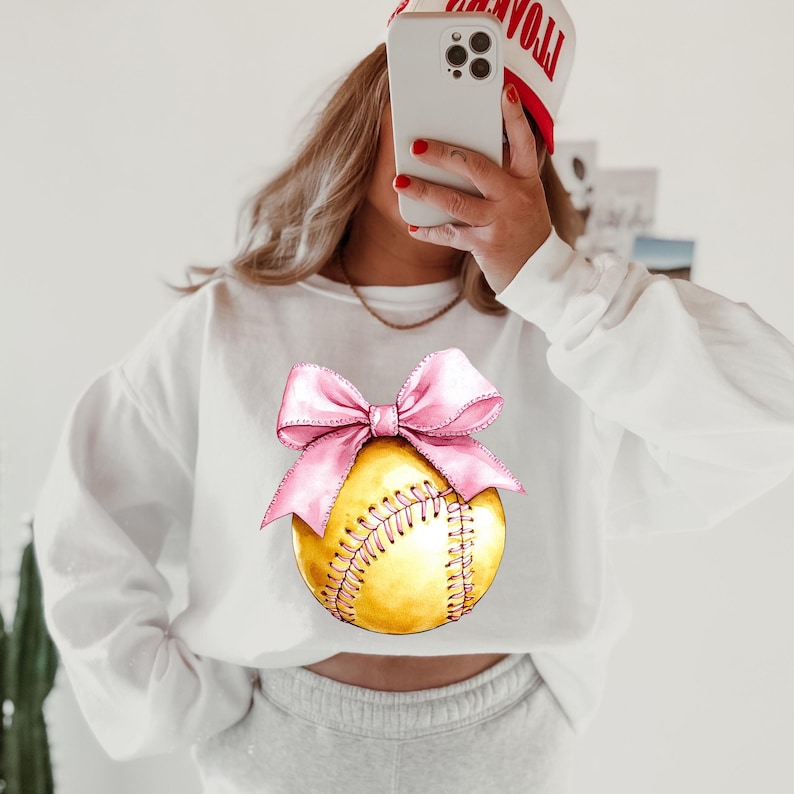 May include: White sweatshirt with a pink bow on a yellow softball graphic.