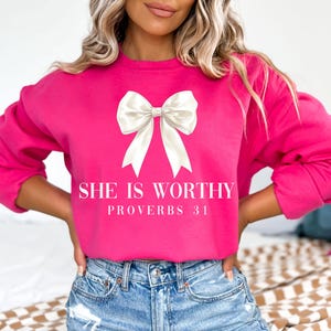 May include: A bright pink sweatshirt with a large white bow graphic. The text "SHE IS WORTHY" and "PROVERBS 31" are printed below the bow in a white font. The sweatshirt is a crew neck style.
