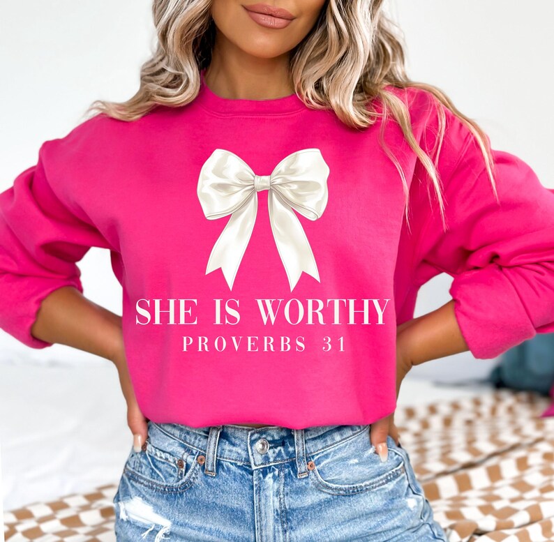 May include: A bright pink sweatshirt with a large white bow graphic. The text "SHE IS WORTHY" and "PROVERBS 31" are printed below the bow in a white font. The sweatshirt is a crew neck style.