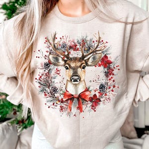 May include: Beige sweatshirt featuring a watercolor illustration of a deer with large antlers, a red bow, and a wreath of pine needles, berries, and flowers. The design is centered on the front of the long-sleeved top, creating a festive holiday theme.