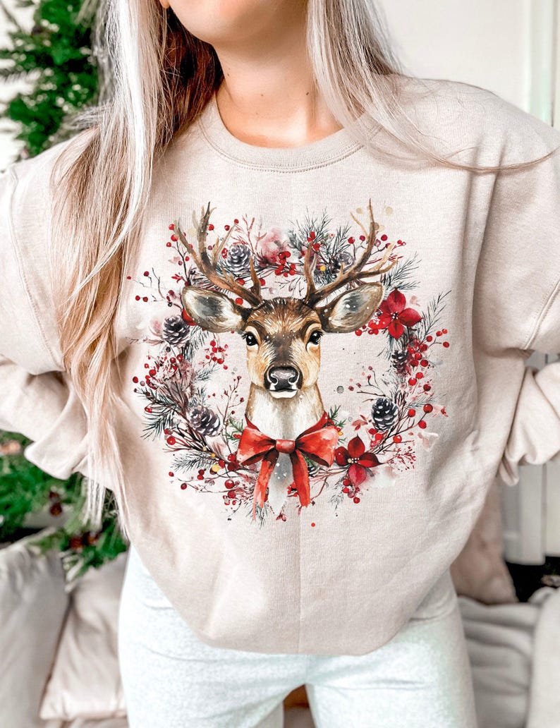 May include: Beige sweatshirt featuring a watercolor illustration of a deer with large antlers, a red bow, and a wreath of pine needles, berries, and flowers. The design is centered on the front of the long-sleeved top, creating a festive holiday theme.