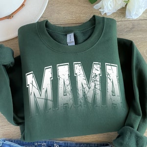 Op de afbeelding: Een donkergroen sweatshirt met een witte, verweerde print die "MAMA" op de voorkant zegt.