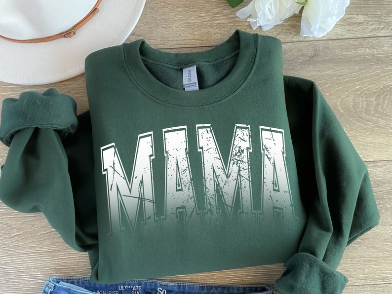 Op de afbeelding: Een donkergroen sweatshirt met een witte, verweerde print die "MAMA" op de voorkant zegt.