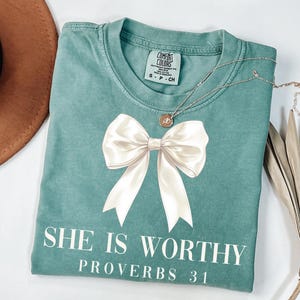 May include: A teal Comfort Colors t-shirt with a large white bow graphic. The text "SHE IS WORTHY PROVERBS 31" is printed below the bow. A silver necklace with a small pendant is draped across the shirt.