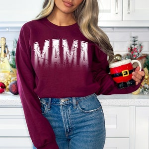Op de afbeelding: Een bordeauxrood sweatshirt met het woord "MAMA" bedrukt in wit met een distressed effect. Het sweatshirt wordt gedragen door een persoon in een keukenomgeving.