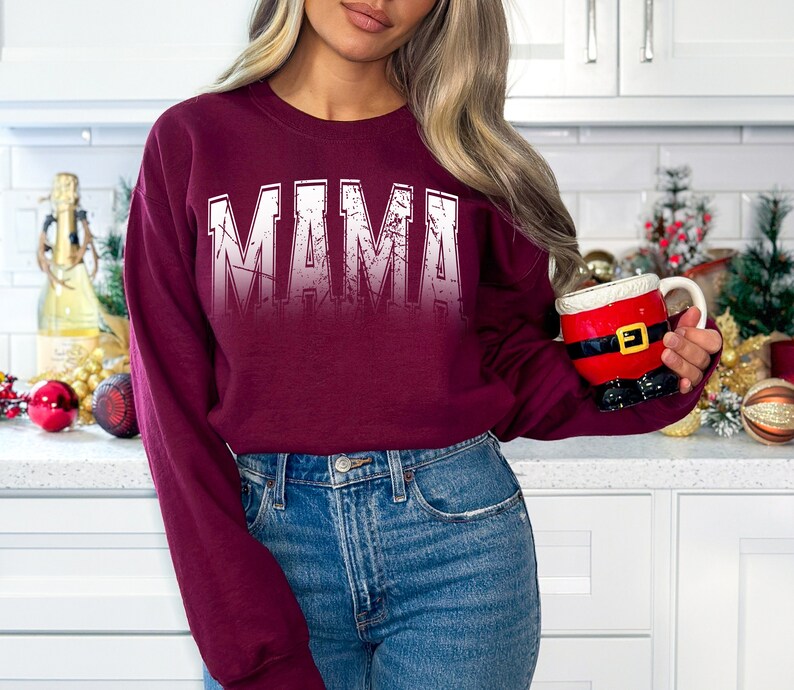 Op de afbeelding: Een bordeauxrood sweatshirt met het woord "MAMA" bedrukt in wit met een distressed effect. Het sweatshirt wordt gedragen door een persoon in een keukenomgeving.