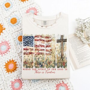 May include: A white t-shirt with a watercolor design of an American flag, flowers, and a wooden cross. The text "Where the Spirit of the Lord is There is Freedom" is printed below the design.