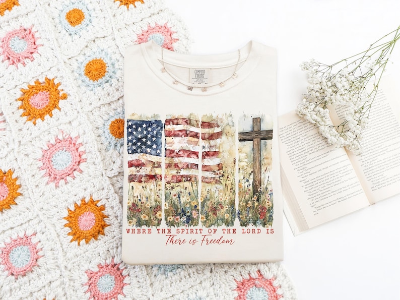 May include: A white t-shirt with a watercolor design of an American flag, flowers, and a wooden cross. The text "Where the Spirit of the Lord is There is Freedom" is printed below the design.