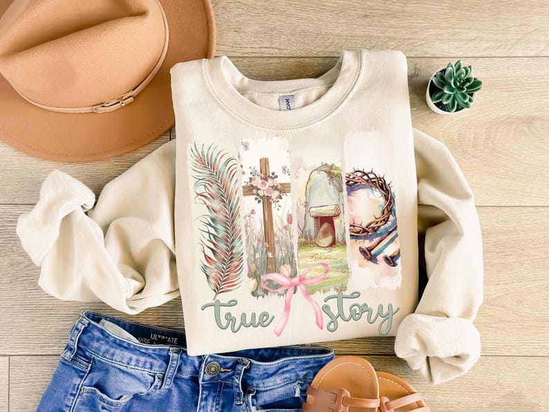 Puede incluir: Una sudadera beige con un dise&ntilde;o gr&aacute;fico que presenta una cruz, una corona de espinas, una tumba y una rama de palma. El texto "true story" est&aacute; impreso debajo del gr&aacute;fico.