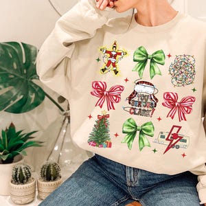 May include: Beige sweatshirt featuring Christmas-themed graphics. The design includes a Santa Claus figure, green and red bows, a Christmas tree, string lights, and the phrase "You Serious Clark!".