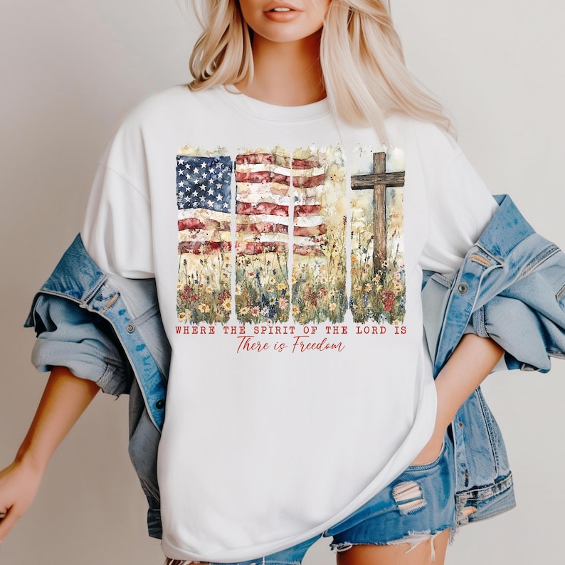May include: White t-shirt with a watercolor design of an American flag, flowers, and a wooden cross. The text "Where the Spirit of the Lord is There is Freedom" is printed below the design.