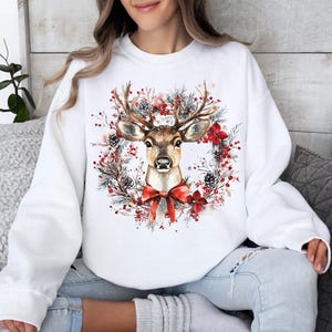 May include: White sweatshirt featuring a watercolor illustration of a deer with antlers, a red bow, and a wreath of pine needles, pine cones, and red flowers. The design is centered on the front of the sweatshirt.
